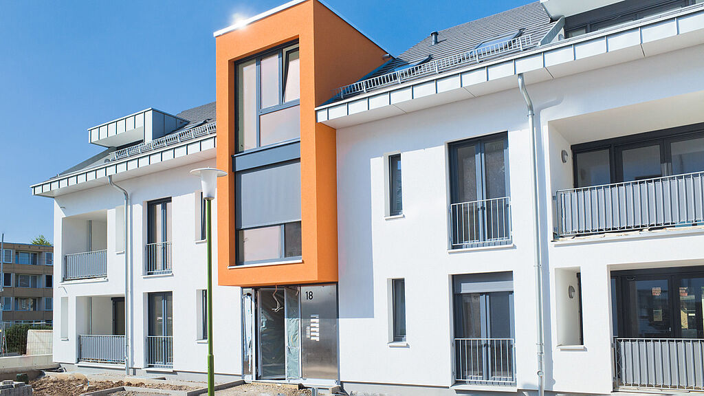 Modernes Mehrfamilienhaus, Naubau, mit weißer Fassade und orangenen Akzent über der Eingangstür